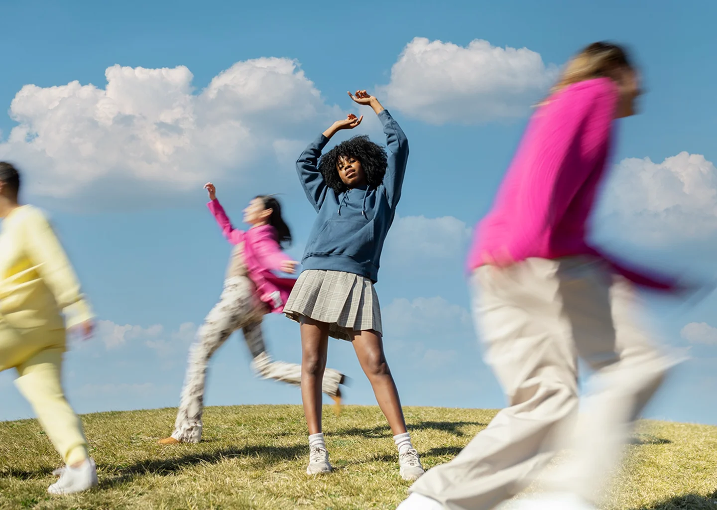 A young woman in a blue hoodie stands on a grassy hill while friends blur past around her under a bright blue sky.